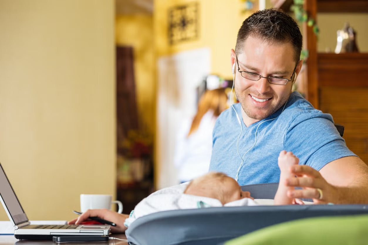 Man working on a laptop at home while holding a baby in a rocker beside him.