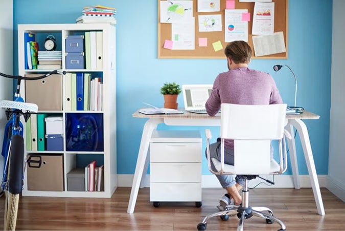 Person sitting at white desk in organized home office with blue walls, bulletin board, and white storage shelving