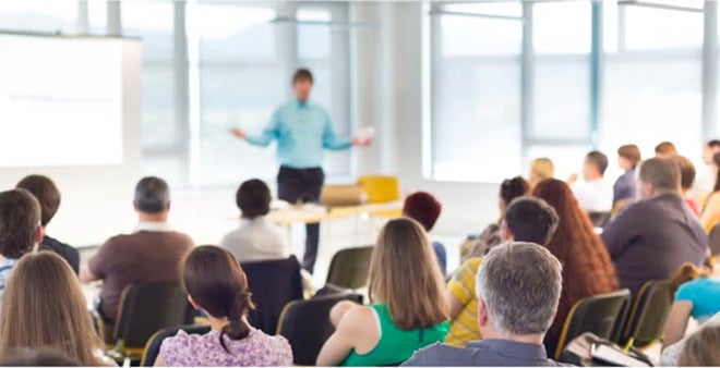 Back view of diverse audience seated and watching male presenter in blue shirt during seminar or workshop in bright modern space