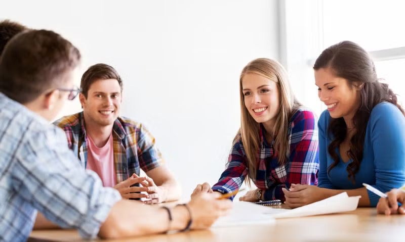 A small group of adults sitting around a table, smiling and talking together, with notebooks and drinks in front of them.