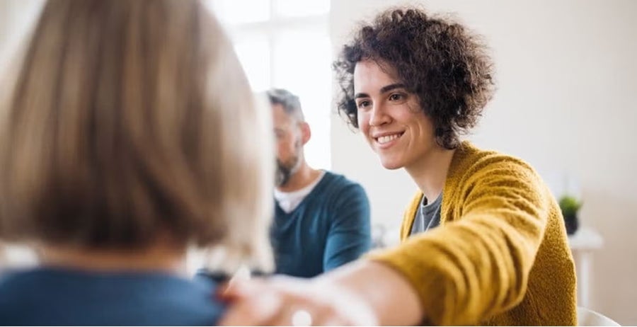 Counselor with curly hair in yellow cardigan smiling warmly during group counseling session with clients