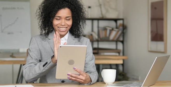 Smiling woman with curly hair in grey blazer waving at tablet in video call with coffee cup and laptop nearby