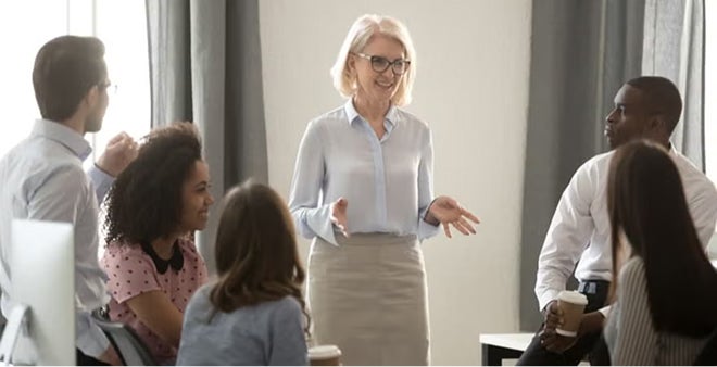 Smiling senior woman with blonde hair and glasses in white blouse speaking with diverse professional team in bright modern office