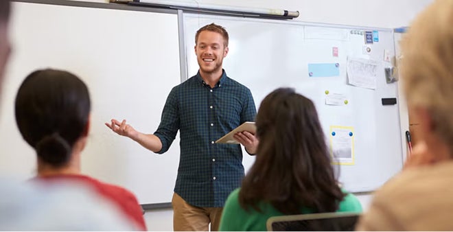 Smiling male instructor in plaid shirt presenting to students in casual classroom training session with whiteboard