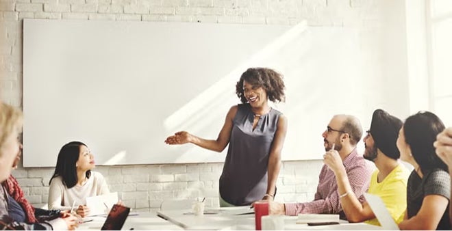 Woman with curly hair presenting to diverse team in modern conference room with whiteboard and collaborative workspace