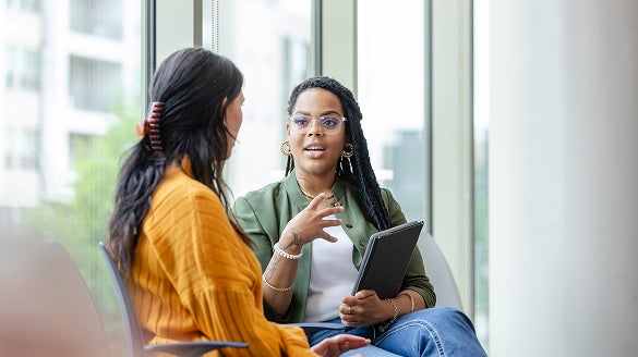 Two women engaged in conversation in a modern office environment, surrounded by desks and office supplies.