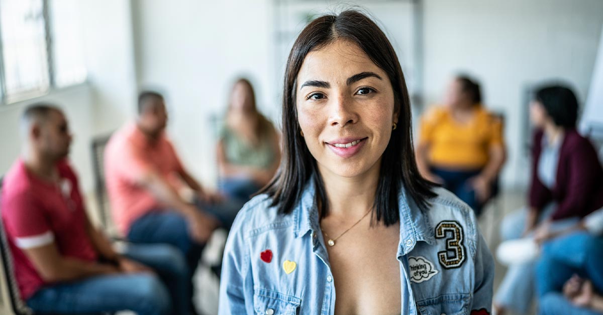 A woman with a warm smile stands in front of a group of people.
