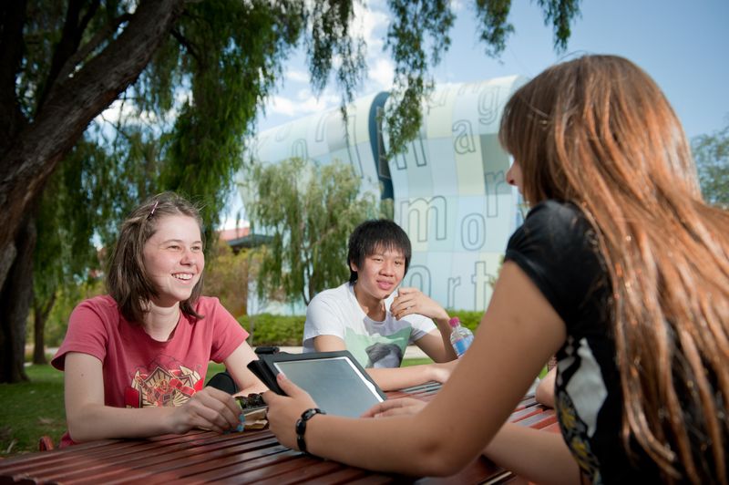 A counsellor and two children sit at a table outside. The counsellor has a tablet device and they are all talking and smiling.