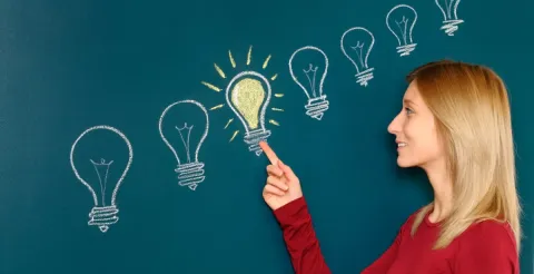 A young woman with long blonde hair in a red sweater stands beside a dark teal wall with a chalkboard effect, pointing to a glowing yellow lightbulb in a sequence of white outline lightbulbs arranged in a rising diagonal line, symbolizing idea generation or innovation.