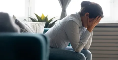Woman sitting on teal couch appearing distressed with hands covering face depicting mental health struggles in bright living room