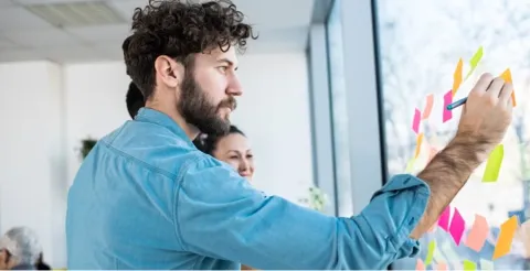 Man with curly hair in blue shirt organizing colorful sticky notes on glass wall during project planning session