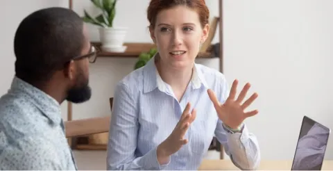 Woman in striped blouse gesturing while speaking to colleague in casual office meeting with plants in background