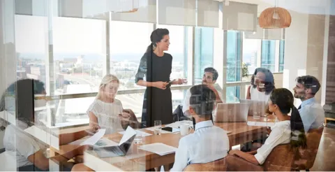 Professional woman in black dress presenting to diverse team at glass conference table in bright modern office with city views