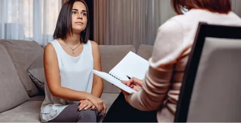 Young woman with dark hair sitting on couch in therapy session with counselor taking notes
