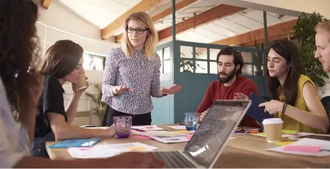 Woman with glasses explaining ideas to diverse team at casual meeting table in modern office with exposed beams