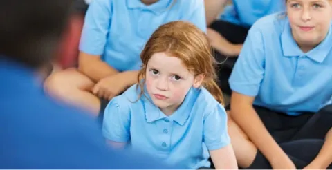 Young girl with red hair and freckles in blue school uniform looking thoughtfully at teacher during class