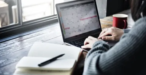 A student’s hands writing in a notebook next to a laptop, phone, and coffee on a tidy desk, representing time management and planning.