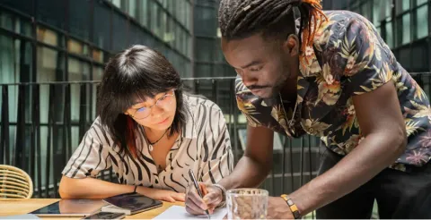 Two diverse university students collaborating over paper and tablet at library table with books in background