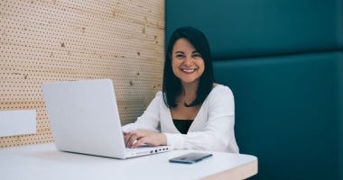 An ECU Online Master of HRM student sits at a table working on her laptop and smiling at the camera.
