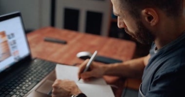 An ECU Online Student is sitting at a desk. They have their laptop and are taking notes with pen and paper.